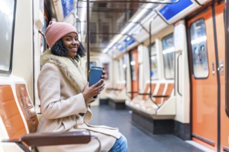 Smiling young black woman wearing winter clothes holding reusable coffee cup while riding subway