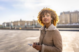 Happy beauty latin young using phone and listening to music standing in the city during sunset