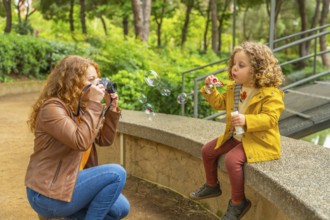 Caucasian adult woman taking a photo with a digital camera of her daughter blowing soap bubbles in