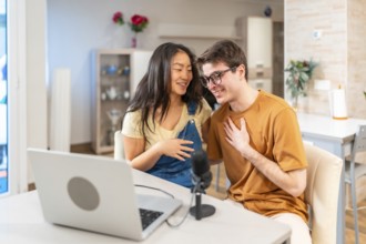 Young multiethnic couple sitting together at home, using a laptop and microphone to record their