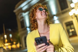 Joyful woman wearing modern sunglasses using mobile app at night in the city