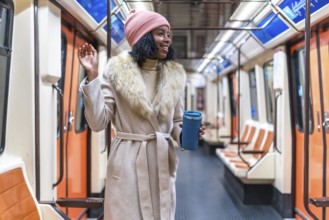 Smiling young black woman wearing winter coat and pink woolen hat holding reusable coffee cup