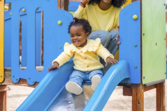 Cute african girl sliding in slide in a colorful playground next to her mother