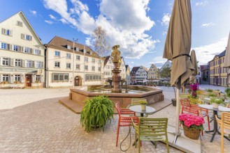 A lively town square with colourful chairs and tables next to a fountain under a clear sky, Weil