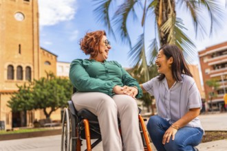 Asian caregiver holding hand with disabled woman in wheelchair outdoors in a beauty city square