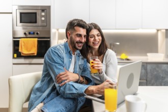 Young couple laughing while watching a movie on a laptop in the kitchen and drinking orange juice
