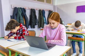 Elementary school students studying at individual desks, girl using laptop, boy writing in