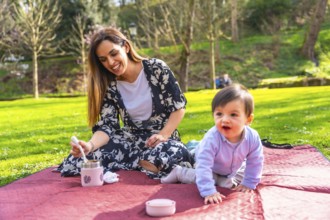 Happy mother preparing a meal from a thermos for her baby during a picnic on a blanket in a sunny