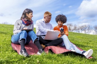 Three students collaborating on a project, using laptop, tablet, and smartphone, enjoying sunny