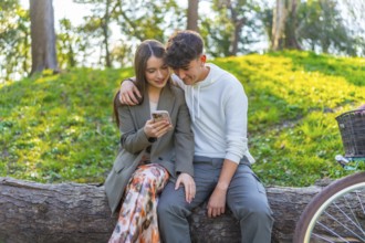 Smiling young couple sitting on a log in a park, looking at a smartphone together, surrounded by