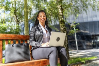 Latin businesswoman smiling at camera while sitting on city bench working with laptop