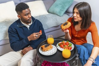 Young multiethnic couple eating pastries and strawberries with orange juice, enjoying breakfast at