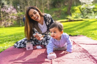 Happy mother feeding her baby solid food with a spoon, sitting on a blanket during a picnic in a