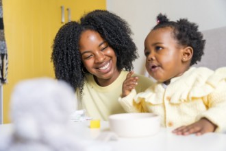 African toddler eating in the living room next to her smiling mother at home