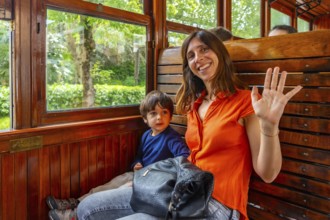 A woman and a child are sitting on a train. The woman is wearing an orange shirt and is waving at