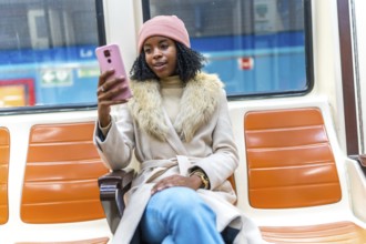 Young black woman wearing winter clothes using her smartphone while traveling on a subway train