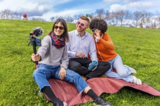 Three content creators are sitting on blanket in park, filming video blog using smartphone with