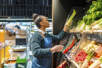 Supermarket employee wearing gloves arranging organic celery on a shelf, surrounded by various