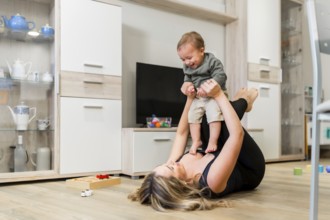 Young mother lying on the floor lifting her baby in the air while playing together in the living