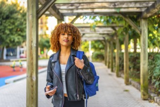 Happy latin female student walking along the city carrying bag and using smartphone