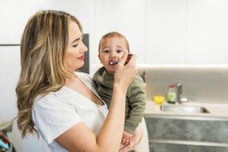 Loving mother feeding her baby son with a spoon in a bright modern kitchen, enjoying a tender