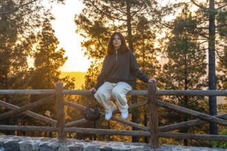 Young woman sits on a wooden fence, taking in the breathtaking sunset over the forested landscape