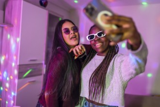Two multi ethnic female friends wearing sunglasses and enjoying a home party, taking selfies with
