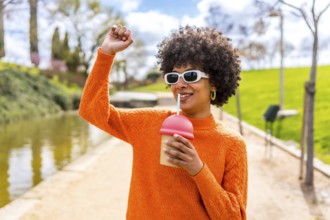 Happy young woman with curly hair and orange sweater, enjoying a smoothie in a park by the river,