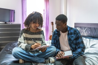 Two focused college students are sitting on a bed, sharing notes and preparing for exams in their