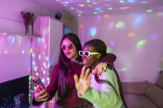 Two teenage girls wearing sunglasses are embracing and taking a selfie with smartphone at a home