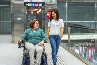Relaxed caucasian woman in wheelchair and asian friend talking and walking along the metro station