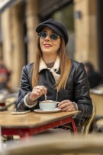Young woman wearing sunglasses and hat stirring cappuccino at a parisian outdoor cafe, enjoying a