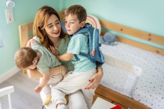 Mother embracing and holding a school boy and baby sitting together in the bedroom before going to