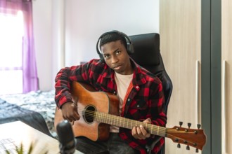 Young man playing acoustic guitar and recording original music in a cozy home studio, immersed in