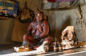 Traditional Himba woman applying ochre paint for body colouring, interior of the first woman's mud