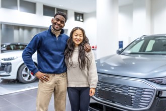 Smiling multiethnic couple buying a new electric vehicle at car dealership, embracing each other