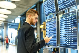 Businessman checking flight information on an airport display while using a smartphone, staying