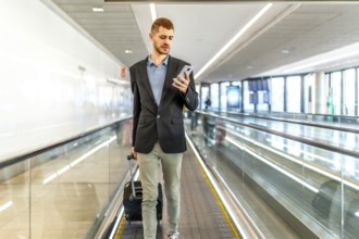 Businessman pulling suitcase and using smartphone while walking on moving walkway at airport