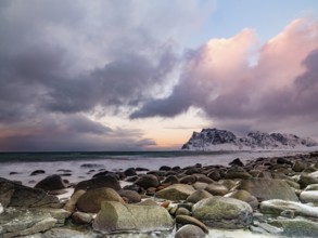 Rounded rocks on Utakleiv beach in a dramatic cloudy atmosphere, snow-capped mountains in the