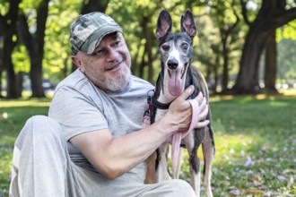 Close-up of a mature bald man smiles while sitting with his greyhound in the park