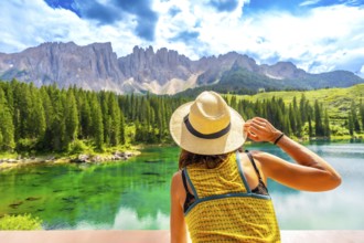 Woman wearing straw hat admiring lake carezza with latemar mountain range in background, dolomites,
