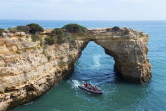 A boat with tourists sails through a natural rock arch over the clear blue sea, Arco de Albandeira,