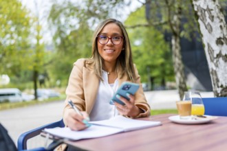Latin woman using phone while writing notes drinking coffee sitting on a tranquil terrace of a
