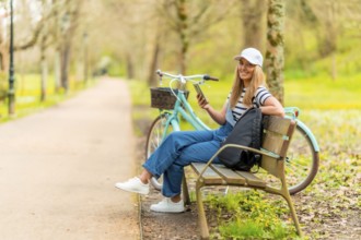 Happy tourist is using her smartphone while resting on a bench in a park with her bicycle next to