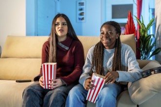 Two teenage girls enjoying a movie night at home, sitting on a comfortable sofa, eating popcorn