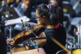 Woman playing the violin in an orchestra on stage, surrounded by music stands and typical concert