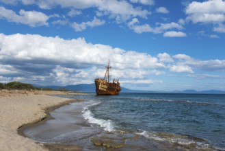 An abandoned, rusty shipwreck lies on a sunny beach with blue sky and clouds, Shipwreck Dimitrios,