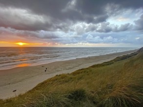 Sunset over the sea under a dramatic, cloudy sky, with people on the beach and grass dunes,