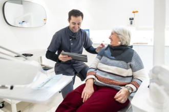 Dentist showing something on tablet to smiling senior woman sitting in dentist chair, during
