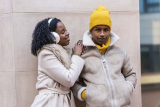 Happy couple dressed in stylish winter clothing, smiling and leaning against a contemporary city